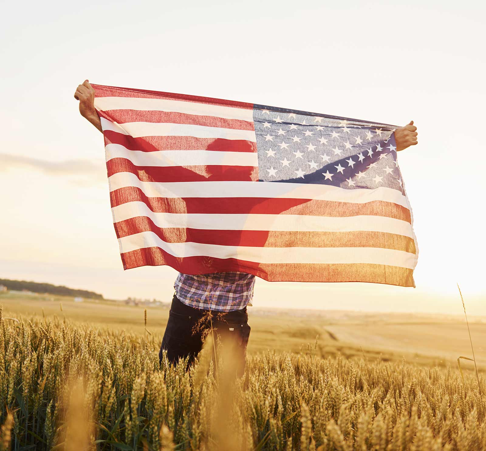 A man holding the American flag high in a wheat field at sunset